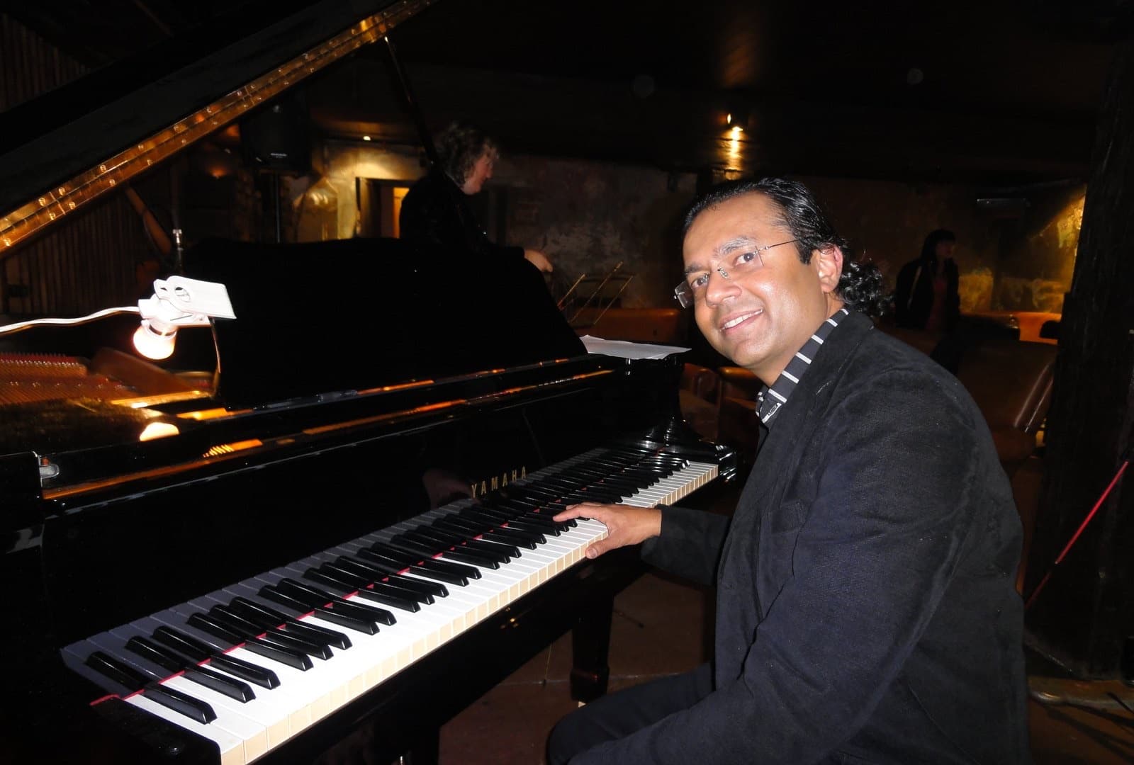 Ben Fernandez seated at a black Yamaha grand piano in a dimly lit jazz club.